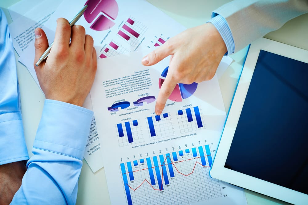 Two professionals analyzing business charts and graphs printed on paper, with one person pointing at the data. A tablet lies on the desk, and various bar and pie charts are visible, reflecting a discussion on business performance.