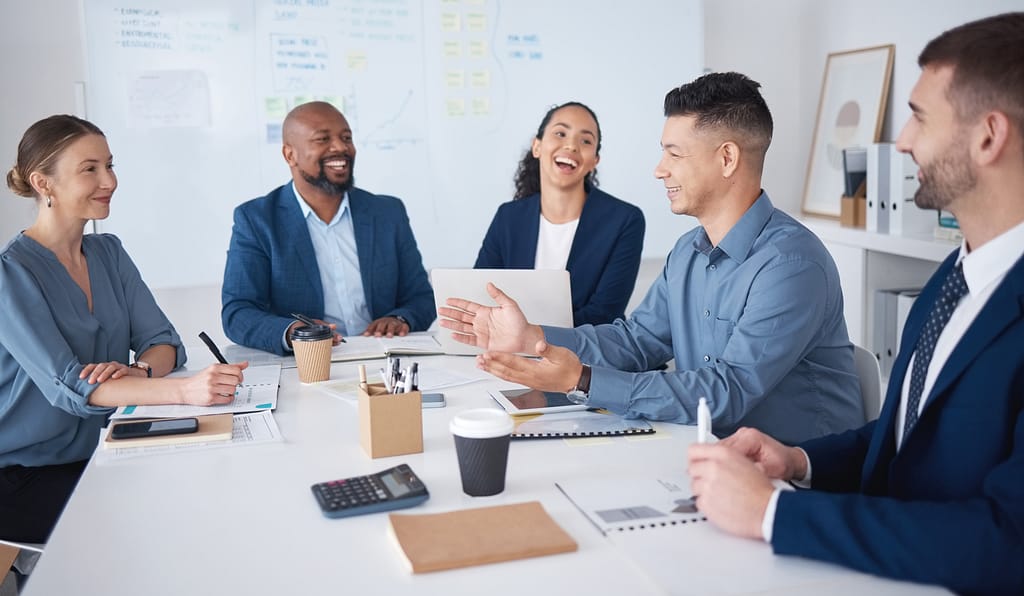 A group of five diverse business professionals engaged in a lively discussion during a meeting. They are seated around a table with notebooks, coffee cups, and office supplies, smiling and interacting in a positive and collaborative atmosphere