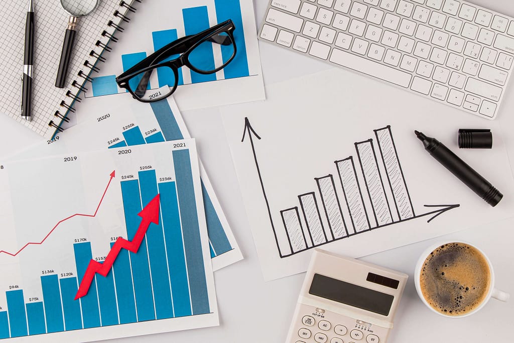 A workspace with various financial charts and graphs, indicating how to increase ecommerce sales over time. The charts show bar graphs with a red upward arrow and hand-drawn bars indicating progress. Nearby are a calculator, a coffee cup, glasses, a keyboard, a magnifying glass, and a notebook.