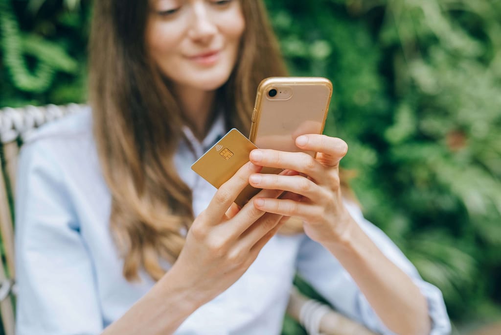 A young woman holding a smartphone in one hand and a credit card in the other, performing an online transaction. She appears to be outdoors with a green, leafy background, dressed casually in a light blue shirt.